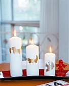 Drei weiße Kerzen mit goldenem Motiv auf einem roten Tablett.