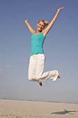 Pretty woman in blue top and white pants jumping on beach with arms outstretched, smiling