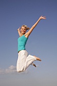 Pretty woman in blue top and white pants jumping on beach with arms outstretched, smiling