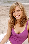 Portrait of beautiful blonde woman wearing purple top sitting on beach, smiling