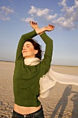 Carefree woman in green sweater, jeans and scarf enjoying sun on beach