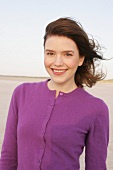 Portrait of pretty woman with windswept hair in purple sweater standing on beach, smiling