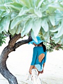 Woman wearing turquoise top and hat, walking under a tree on beach
