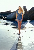 Beautiful blonde woman wearing blue blouse, skirt and boots walking on beach, smiling