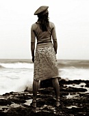 Rear view of woman wearing knitted sweater, chiffon skirt and cap looking at sea on beach