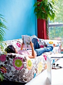 Woman reading book while lying on floral patterned sofa against blue wall