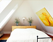 Bedroom with white bed under sloping ceiling and framed picture with floral motif