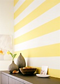 Brown sideboard with vases and bowl in front of yellow and white striped wall