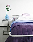Close-up of bed with purple blanket and side table with blue flower vase