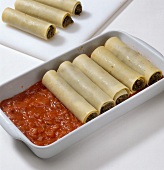 Rolls of stuffed pasta dough with sauce on tray while preparing cannelloni, step 2