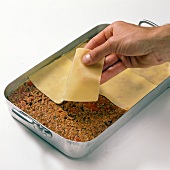 Dough sheet being spread on meat sauce in baking tray