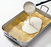 Bechamel sauce being poured on layers of dough sheet and meat sauce in baking tray