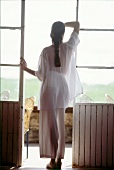 Rear view of brunette woman with pigtail wearing white nightwear, standing at window