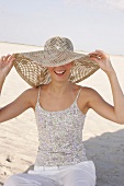 Happy woman wearing large straw hat sitting on sand at beach, smiling