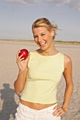 Frau am Strand hält einen roten Apfel in der Hand, lacht