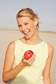 Woman holding red apple standing on beach, laughing
