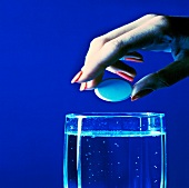 Close-up of woman's hand about to drop tablet in a glass of water against blue background