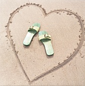 In Sand gemaltes Herz mit zwei Sandaletten in der Mitte
