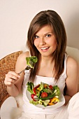 Portrait of woman with long hair holding bowl of salad and eating cucumber, smiling