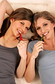 Two women lying on bed and biting green hot chilli pepper and red hot chilli pepper