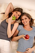 Two young woman lying while holding green and red apple, smiling