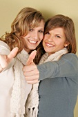 Two beautiful woman in sweater and scarf standing side by side showing thumbs up, smiling