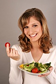 Portrait of young woman holding salad plate in one hand and tomato in another, smiling