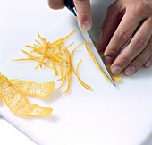 Close-up of hand cutting thin strips of orange peel on white background