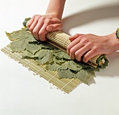 Close-up of hand rolling bamboo mat stuffed with rice on grape leaves, step 3