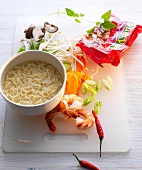 Noodles in bowl with shrimp, mushrooms, carrots and chilli on chopping board