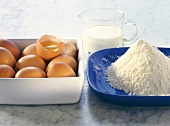 Cup of milk and heap of flour and eggs on plate