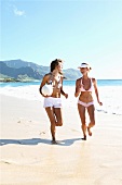 Two cheerful women wearing bikini running on beach with a ball, smiling