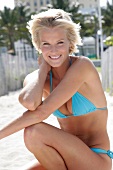 Beautiful blonde woman with windswept hair in light blue bikini sitting on sand, smiling