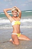 Portrait of pretty blonde woman wearing flower pattern bikini kneeling on beach, smiling