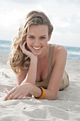 Portrait of beautiful blonde woman wearing brown top lying on beach, smiling