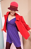 Cheerful young woman wearing purple dress, red jacket and hat looking down and smiling