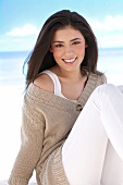 Portrait of pretty woman in beige sweater and white pants sitting on the beach, smiling