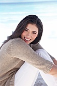 Portrait of pretty woman in gray sweater and white pants sitting on beach, smiling