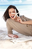 Happy woman wearing beige sweater lying on a blanket on sand holding starfish, winking