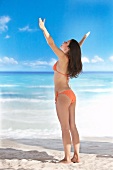Woman in orange bikini raising her arms and standing on beach