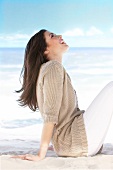 Side view of woman in beige cardigan looking up and sitting on beach, laughing