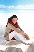 Portrait of woman in beige cardigan and red scarf, sitting on beach and smiling