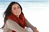 Frau in  Strickjacke, beige, rot, Schal sitzt am Strand, Sand, Meer