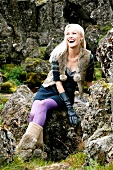 Happy blonde woman wearing fur vest and purple tights, sitting on stone and laughing