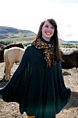 Happy brunette woman wearing poncho and scarf standing in pasture and laughing