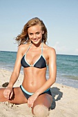 Portrait of beautiful blonde woman wearing gray bikini sitting on beach, smiling