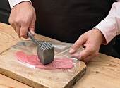 Close-up of hands patting meat with meat tenderizer for preparation of schnitzel, step 1