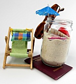 Urlaubsutensilien in einem Glas voll Sand daneben ein Liegestuhl
