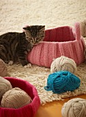 A striped cat next to a pink knitted basket and various different coloured balls of wool on a flocked rug