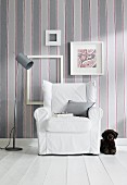 Dog lying next to armchair with white loose cover against striped wallpaper with picture frames